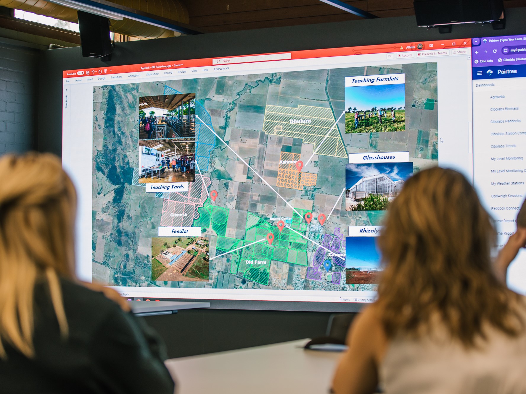 Image depicts two women in the foreground, over their shoulders in the background is a large digital screen showing a farm