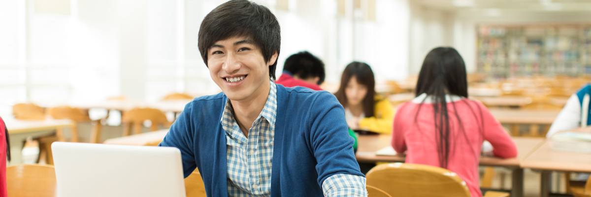 Person sitting at a desk in a library or study area, working on a laptop. Several other people are seated at tables in the background, with bookshelves visible.
