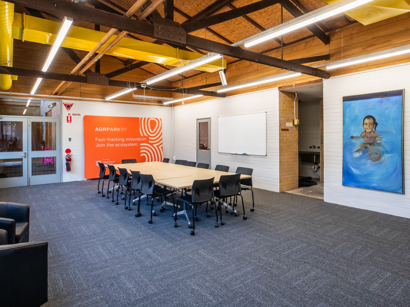 Image depicts a innovation space, a large table surrounded by 15 black chairs, blue carpet, a large blue painting on the wall